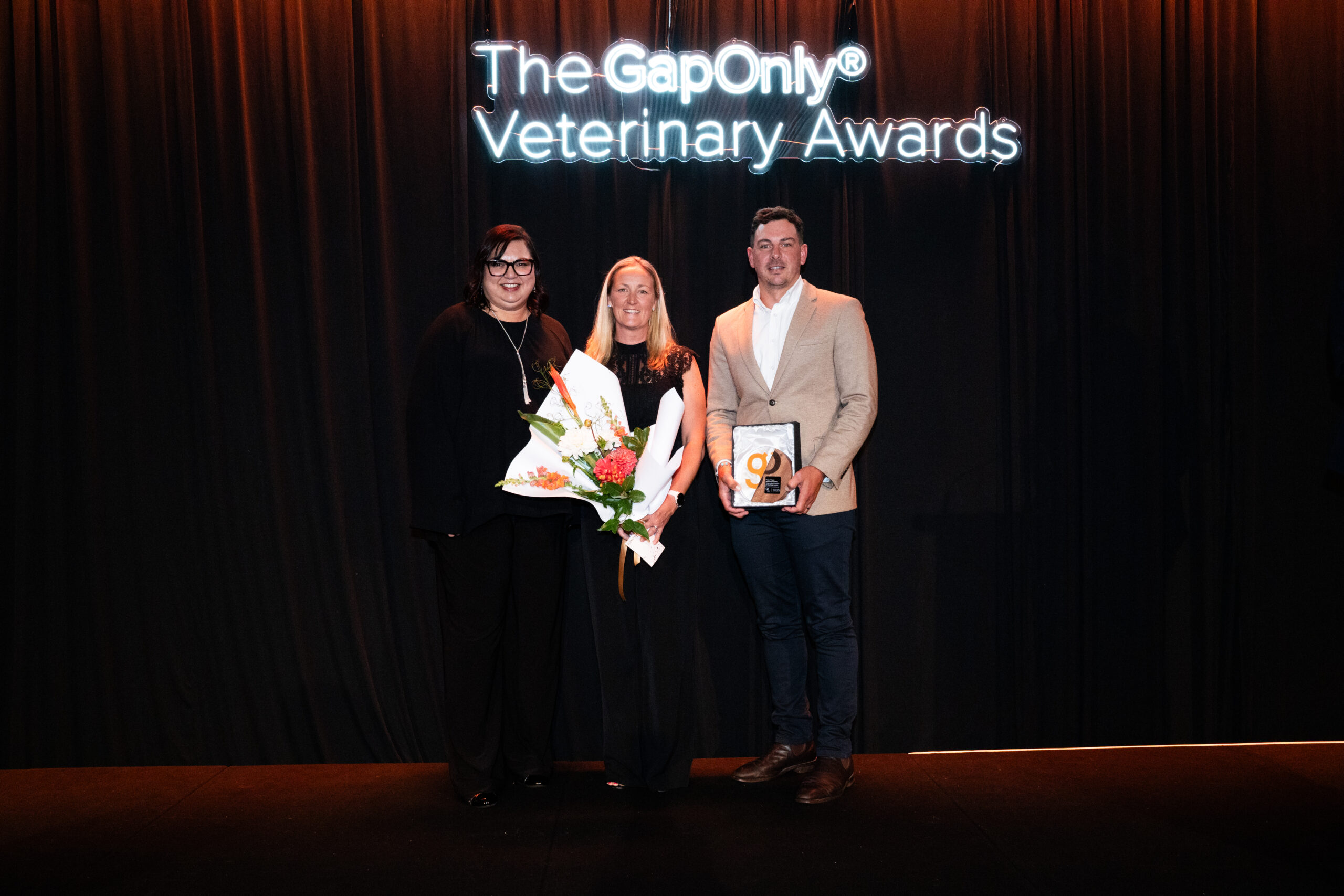 Three individuals stand on stage at the GapOnly® Veterinary Awards, posing for a photo. The person on the left is dressed in black and wearing glasses, the middle individual is holding a bouquet of flowers, and the person on the right is holding an award plaque. A neon-lit event sign is visible in the background.